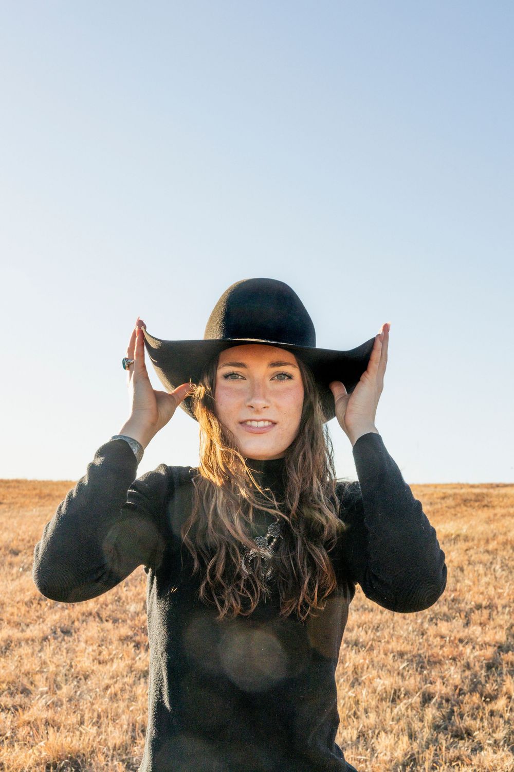 Person in black sweater and wide-brimmed hat standing in golden field during sunset with hands adjusting hat.