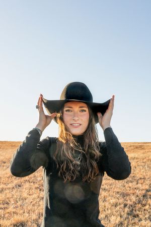 Person in black sweater and wide-brimmed hat standing in golden field during sunset with hands adjusting hat.