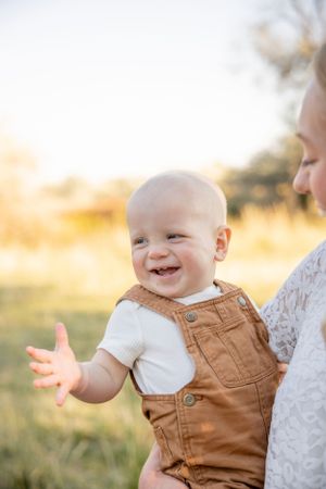 Baby in brown overalls laughing outdoors during golden hour.