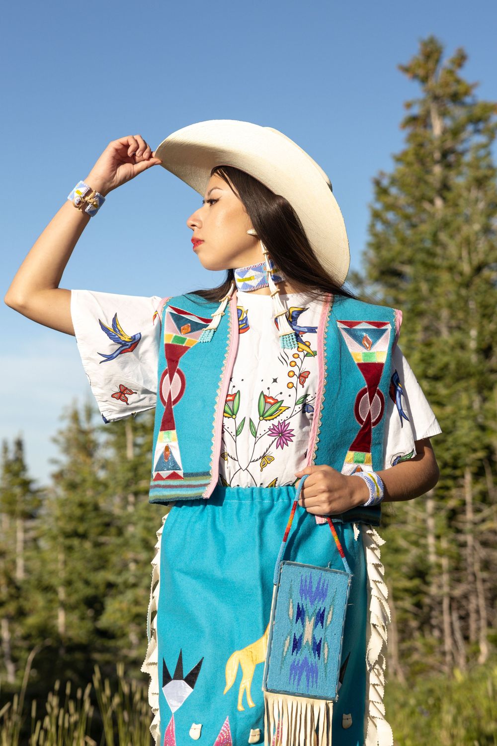 Indigenous traditional dress with turquoise fabric, beading and white cowboy hat against evergreen forest background.