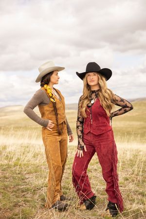 Two friends in western attire including cowboy hats, corduroy overalls and vests pose together in a grassy field.