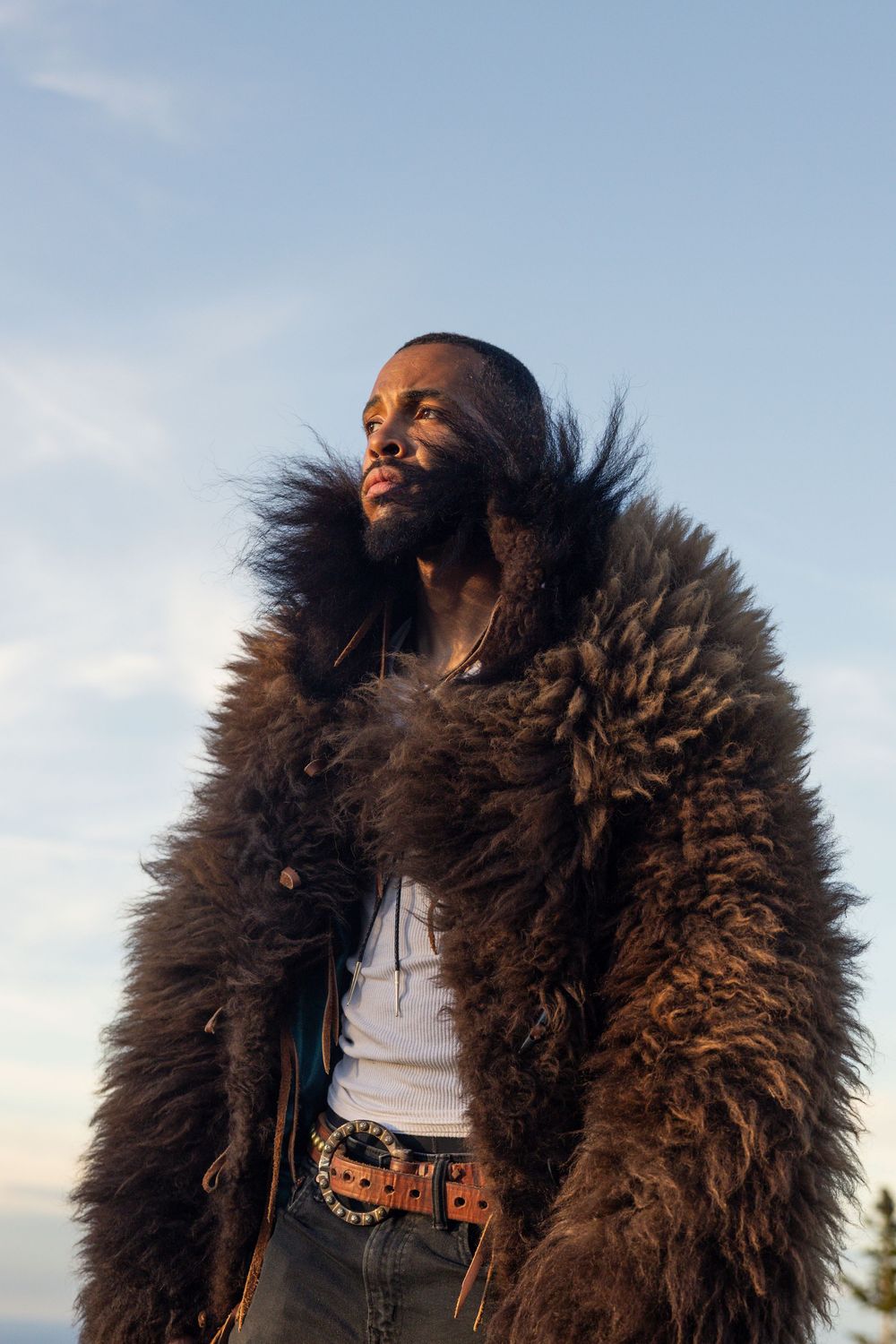 Person wearing a luxurious brown fur coat and white tank top poses against a sunset sky.