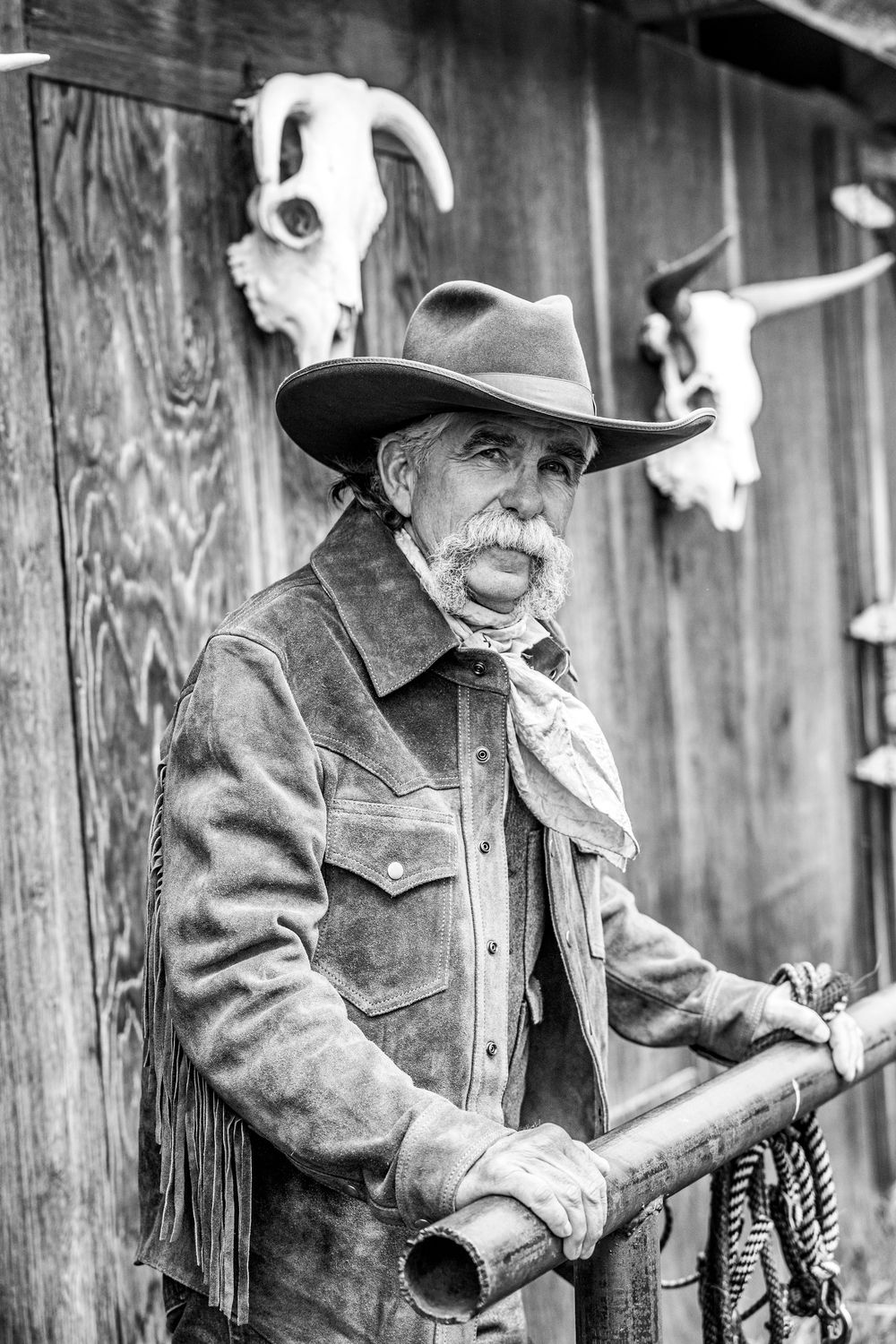 Weathered cowboy in traditional attire poses with animal skulls in black and white portrait.