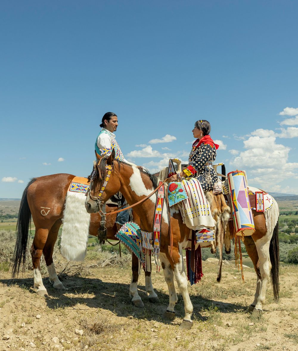 Two Native American riders on decorated paint horses stand on a sunlit plains landscape under a blue sky.