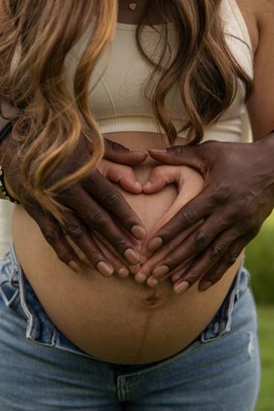 Close up of light and dark skinned hands forming a heart shape over a pregnant belly while wearing jeans and crop top.