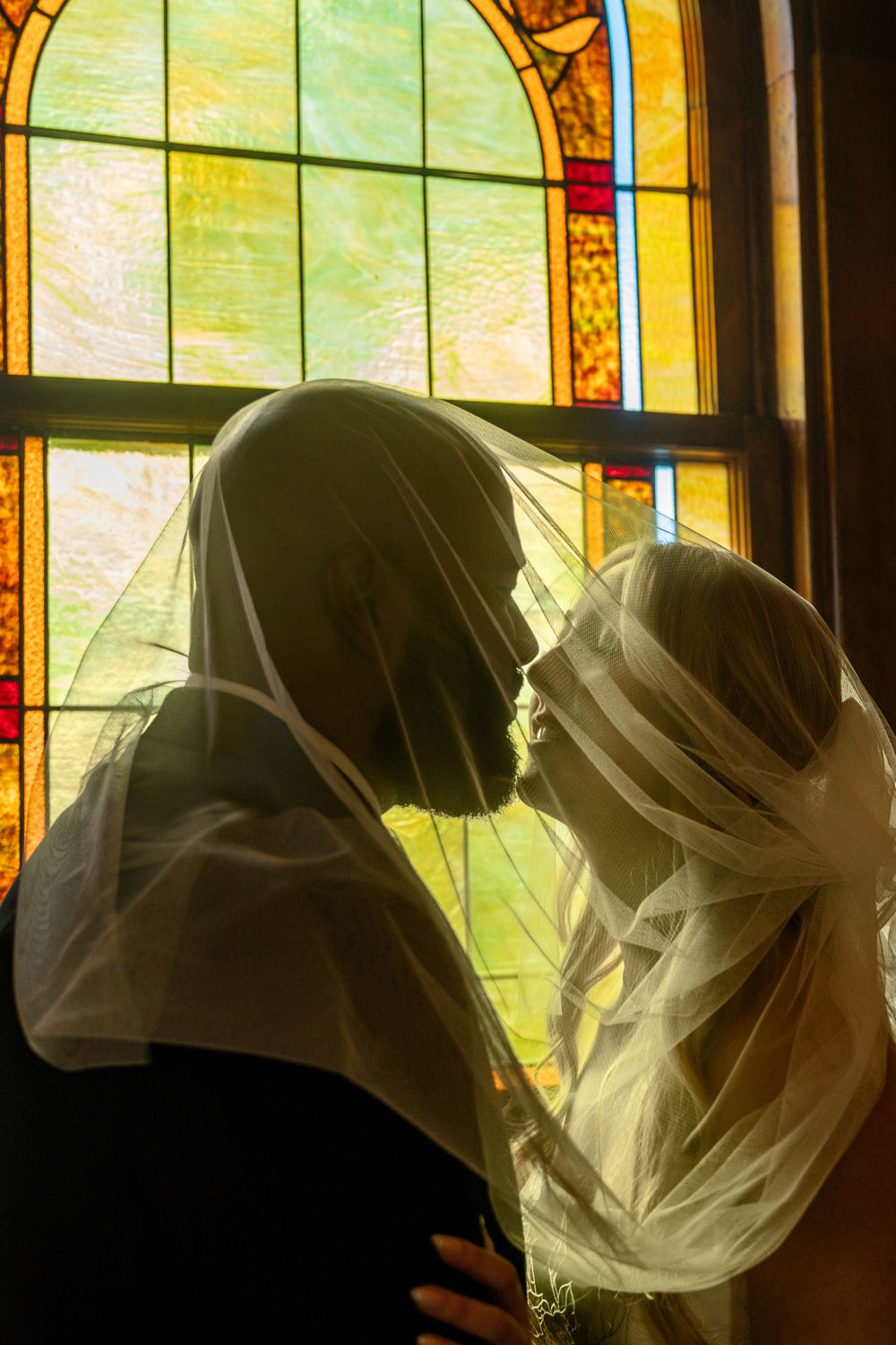 Bride and groom sharing a kiss under a veil before a colorful stained glass church window.