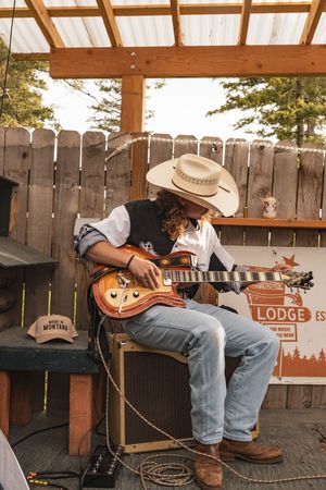 Musicians performing with guitars on rustic wooden patio with lodge sign and warm lighting at sunset.