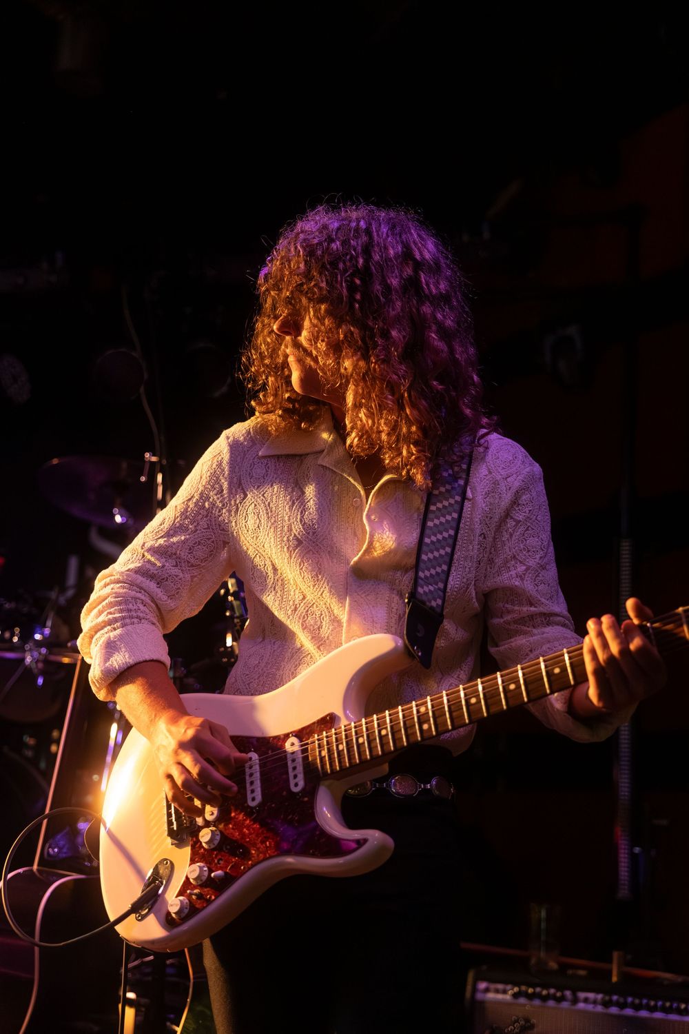 Curly-haired guitarist performing on stage with a white electric guitar under purple stage lighting.