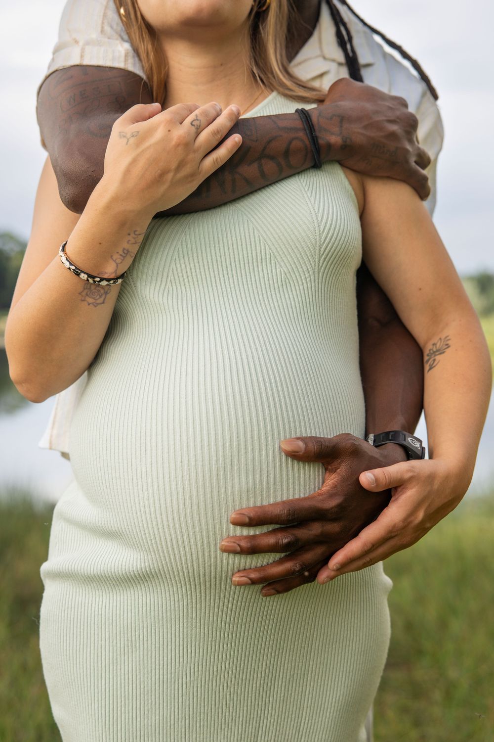 Close up of hands gently holding a pregnant belly in a pale green ribbed dress outdoors.