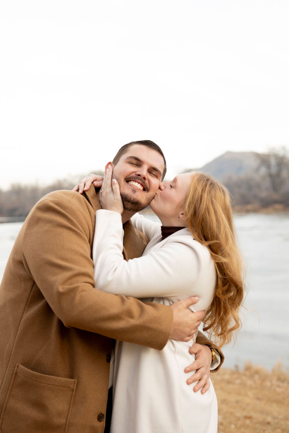 Couple shares affectionate moment by lakeside wearing winter coats.