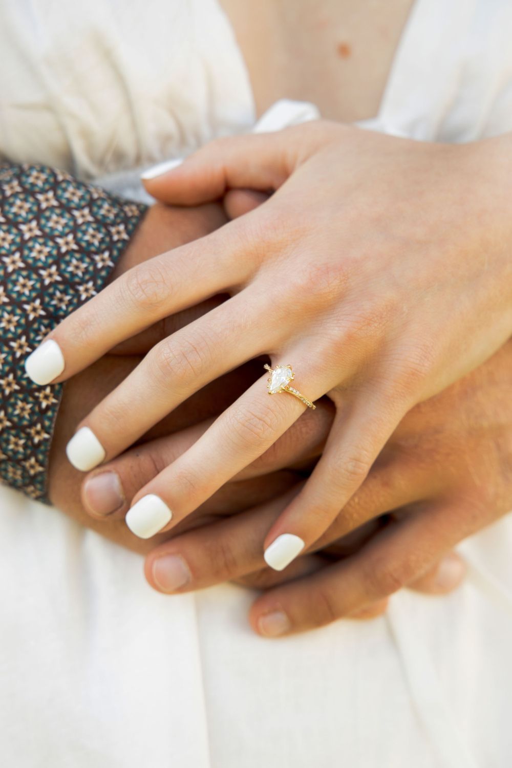 Close-up of hands with white manicured nails and engagement ring against floral fabric.