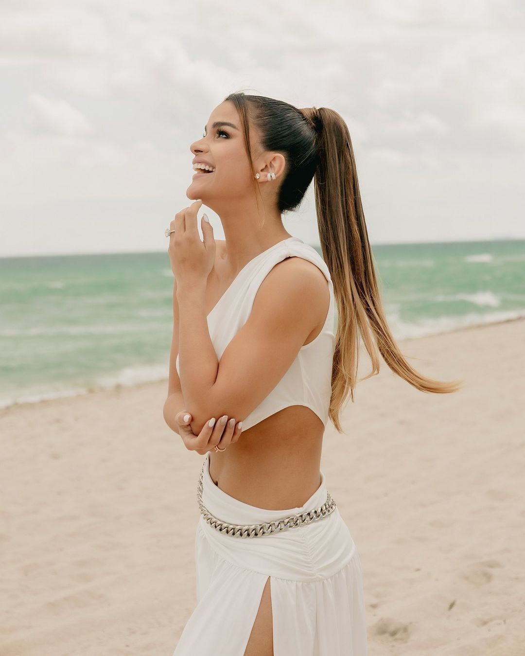Woman in white beachwear smiling while posing on sandy beach with ocean background.