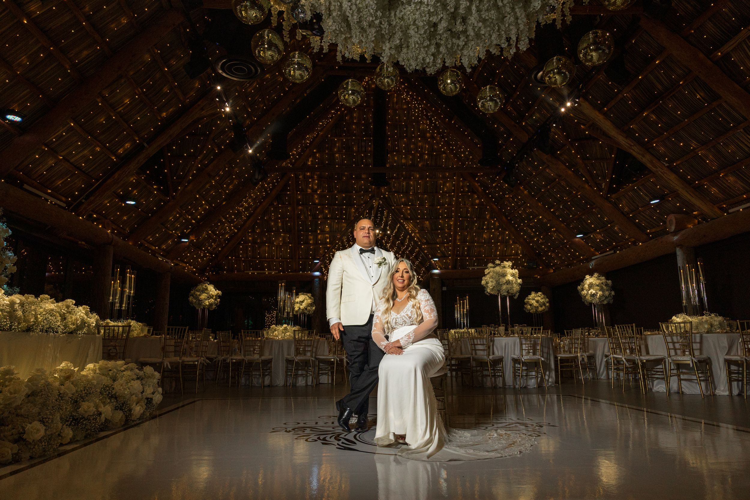 Bride and groom pose elegantly on a dance floor in a stunning barn wedding venue adorned with warm lights and floral arrangements.