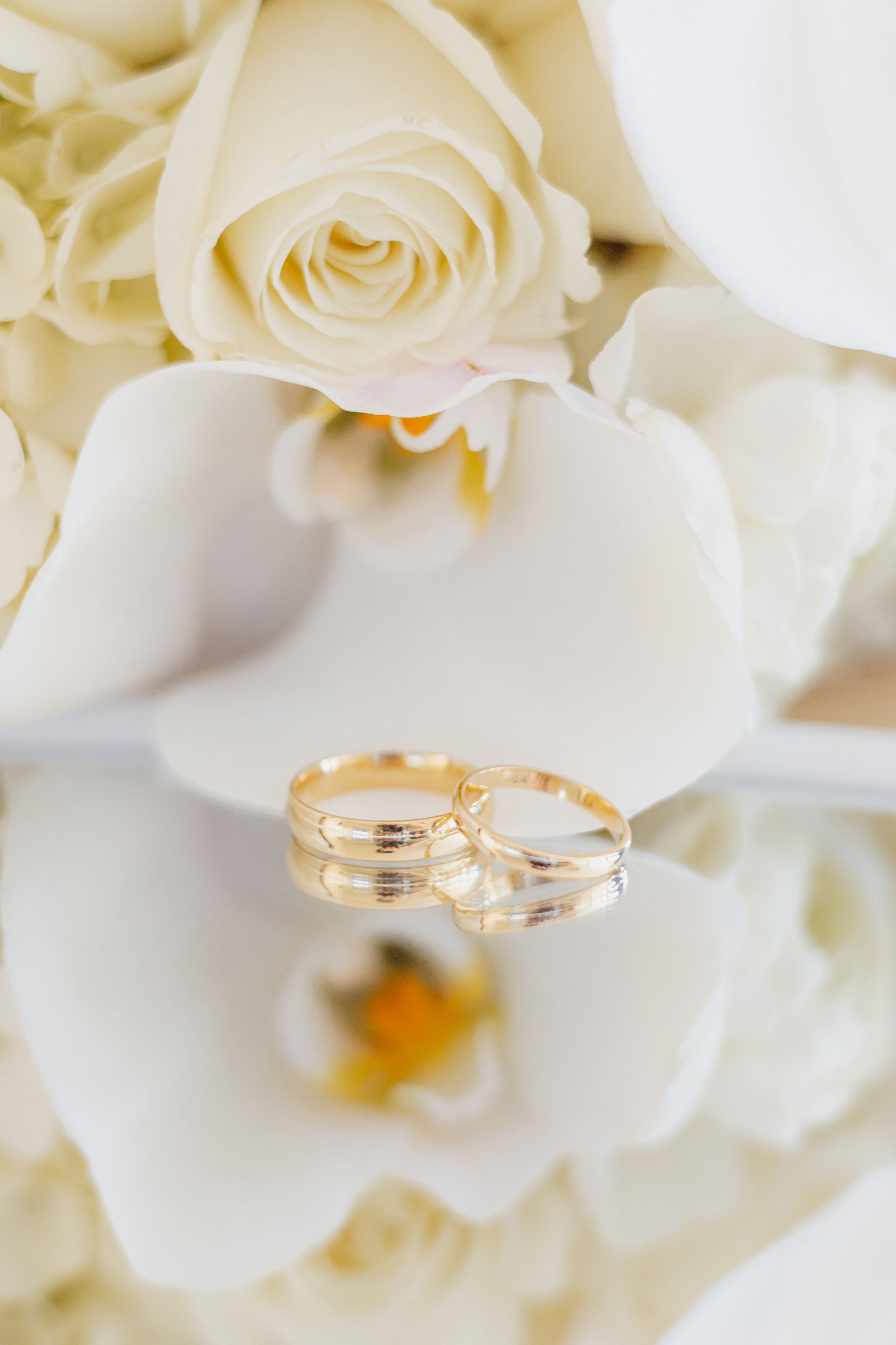Wedding rings displayed on white orchid and rose petals in soft lighting.