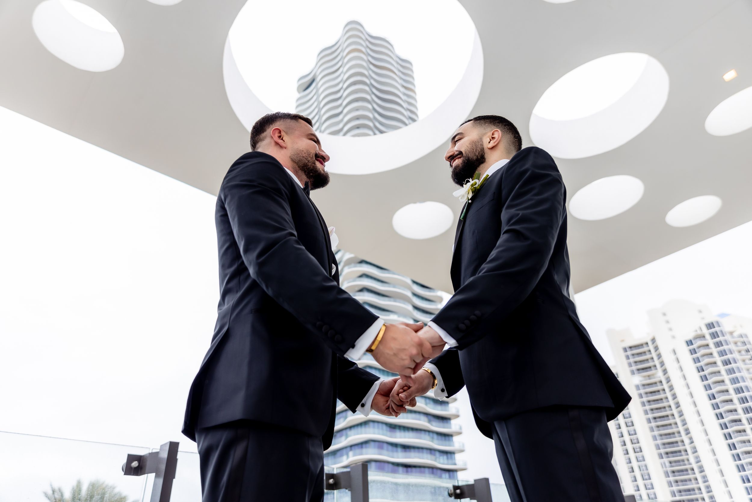 Two businessmen in black suits shaking hands outdoors with modern skyscrapers in the background.