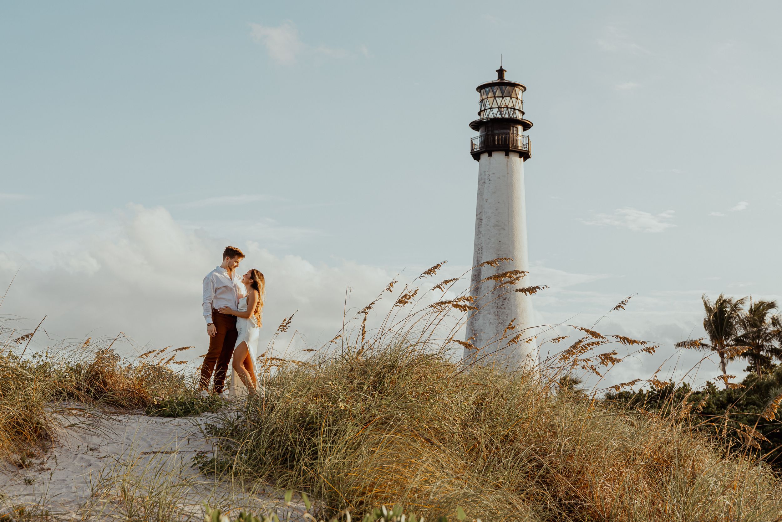 Couple embracing on sandy dunes near a tall white lighthouse on a sunny beach day.