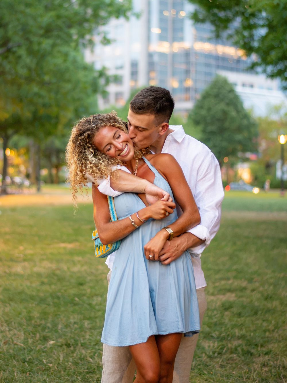 Couple embracing in a park, man kissing woman's cheek, she smiles in blue dress, city buildings in background.