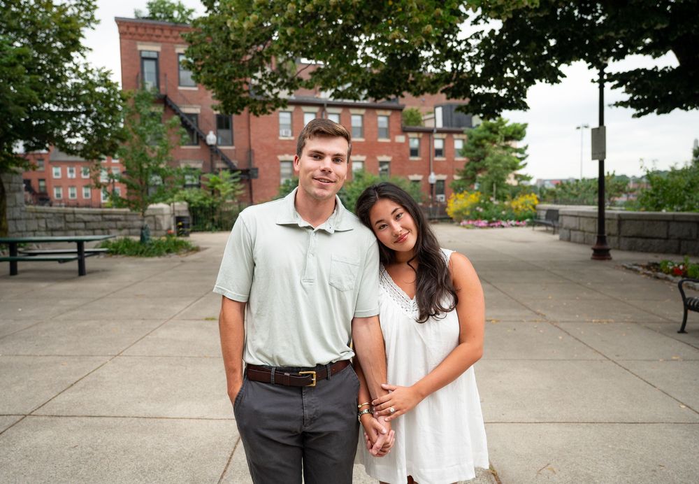 Young couple posing together outdoors in a urban park setting with brick buildings in the background.
