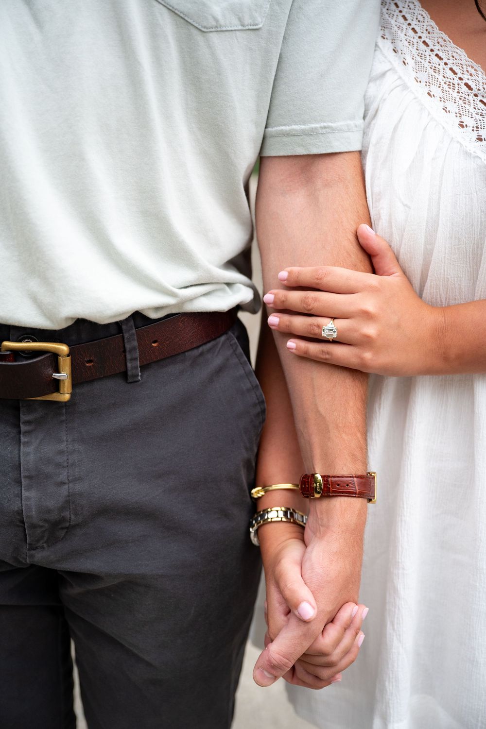 Close-up of couple holding hands, woman wearing engagement ring and bracelets, man in gray pants with brown leather belt.
