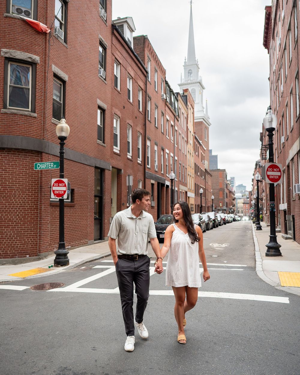 Couple holding hands walking across a street in Boston's North End with brick buildings and church steeple backdrop.