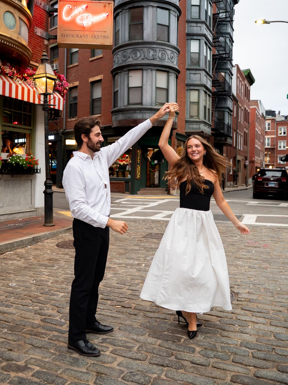 Couple dancing on cobblestone street in Boston, woman in black top and white skirt, man in white shirt and black pants.