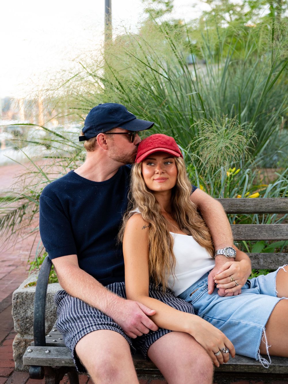 Couple sitting on a bench outdoors, man in navy cap kissing woman's cheek, lush greenery background.