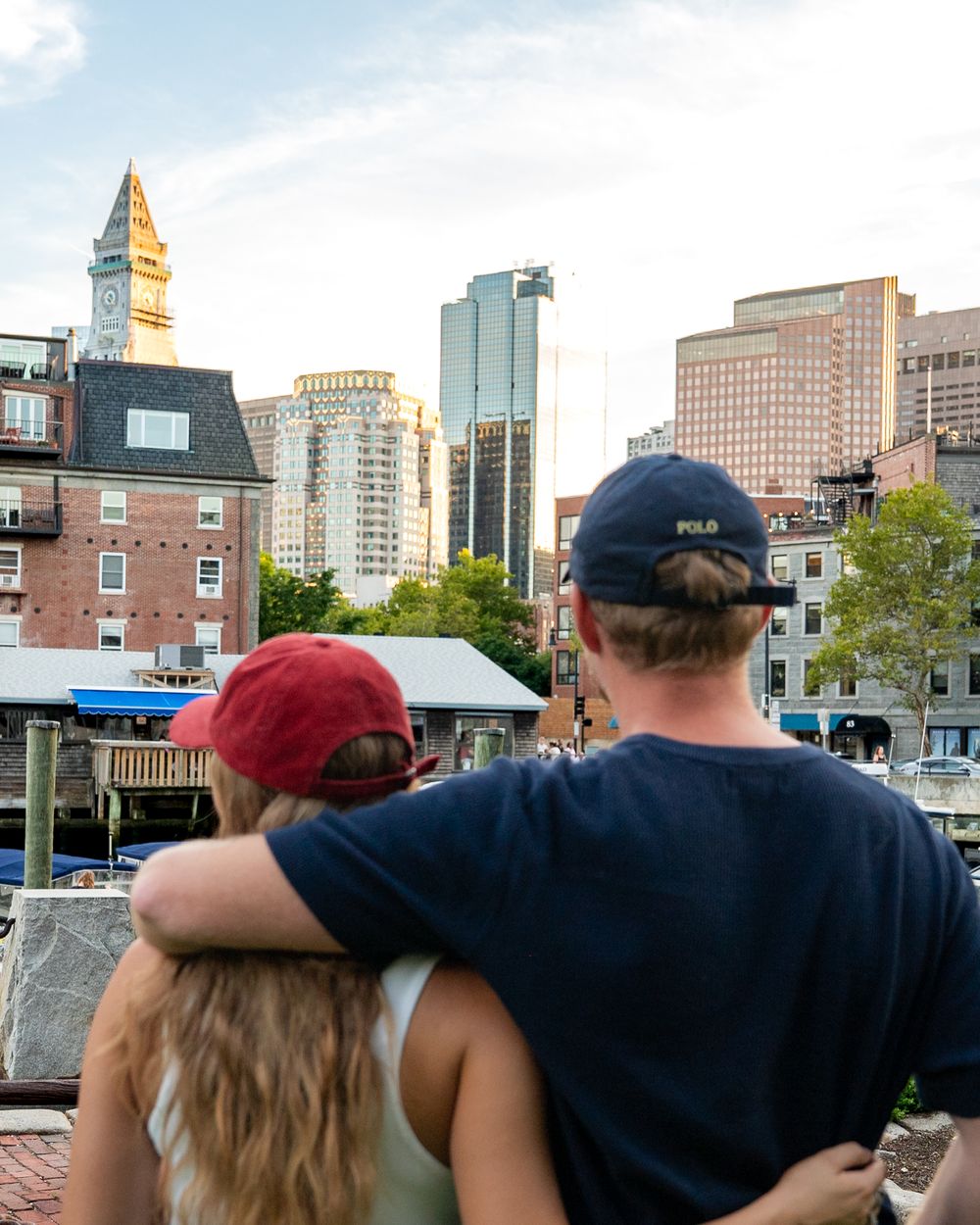 Couple with arms around each other wearing baseball caps admiring Boston city skyline from waterfront.