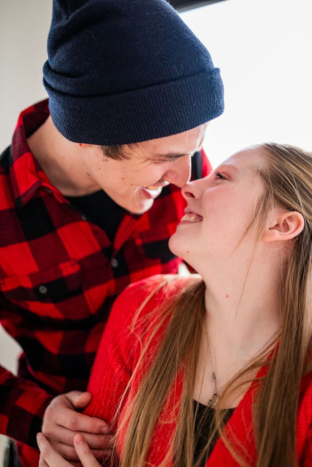 A couple in red and plaid share an intimate laughing moment by a window.
