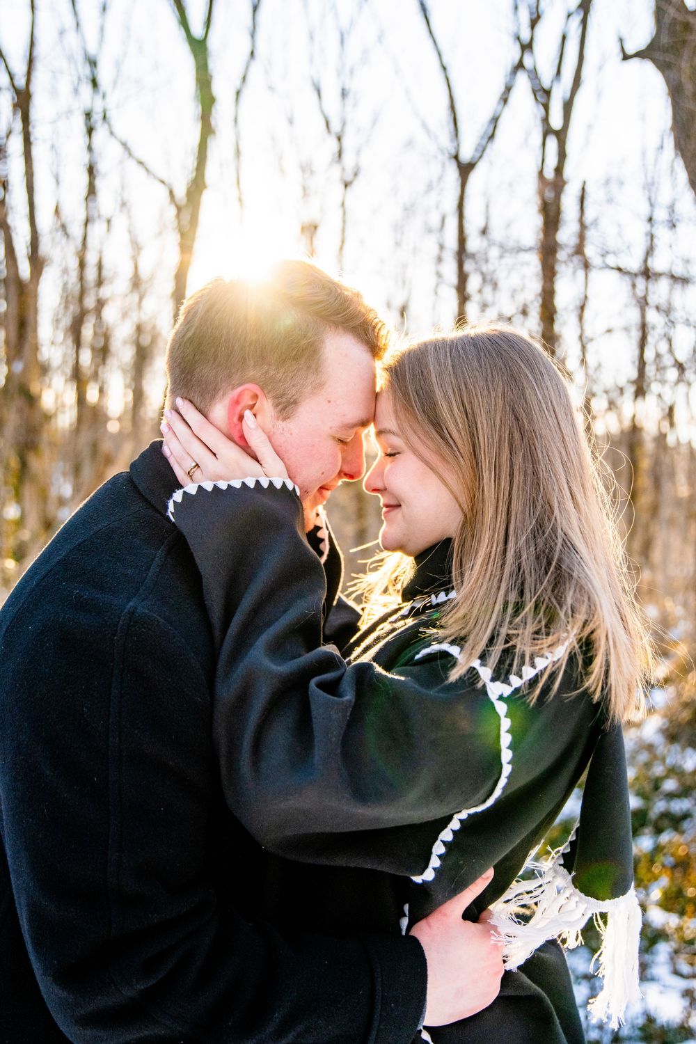 A romantic winter moment captured in the woods as sunlight streams through bare trees in the background.