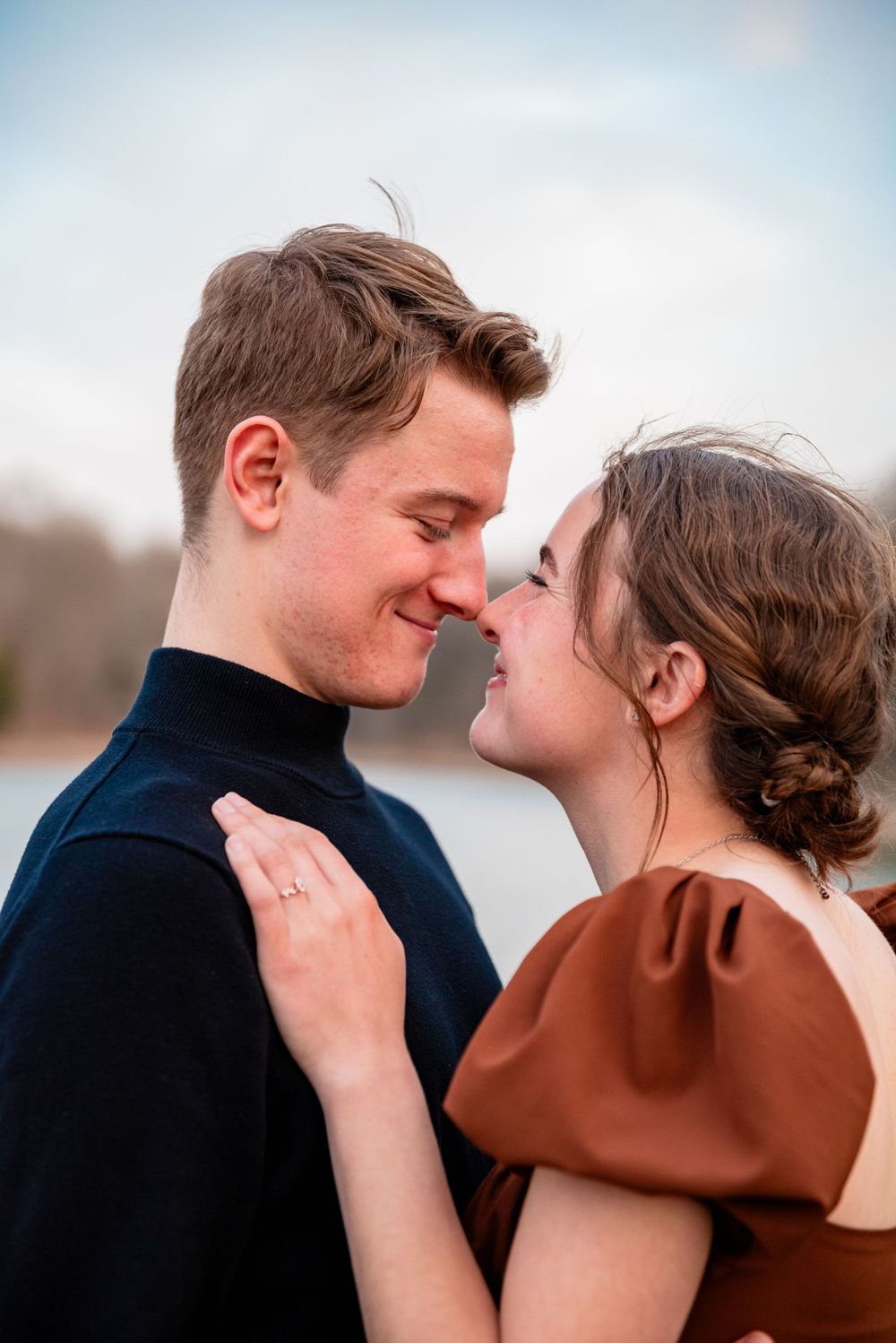 Intimate close-up portrait of couple in black and brown attire against soft sky background.