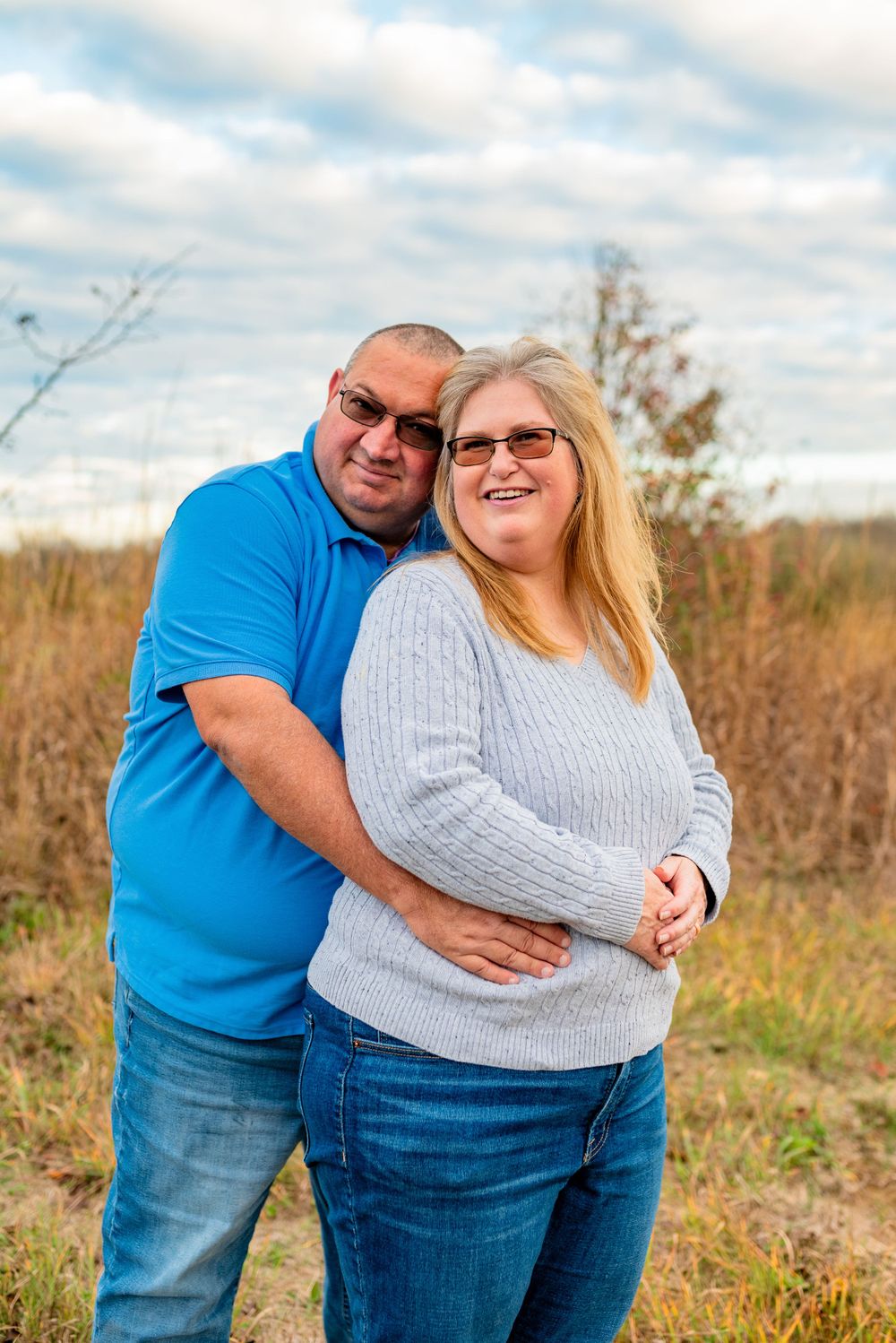 A couple embraces while smiling outdoors in a field on a cloudy day.