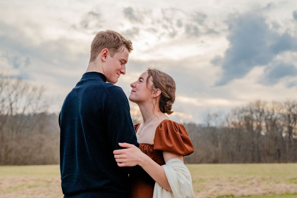 Couple sharing intimate moment outdoors with dramatic sky and trees in background.