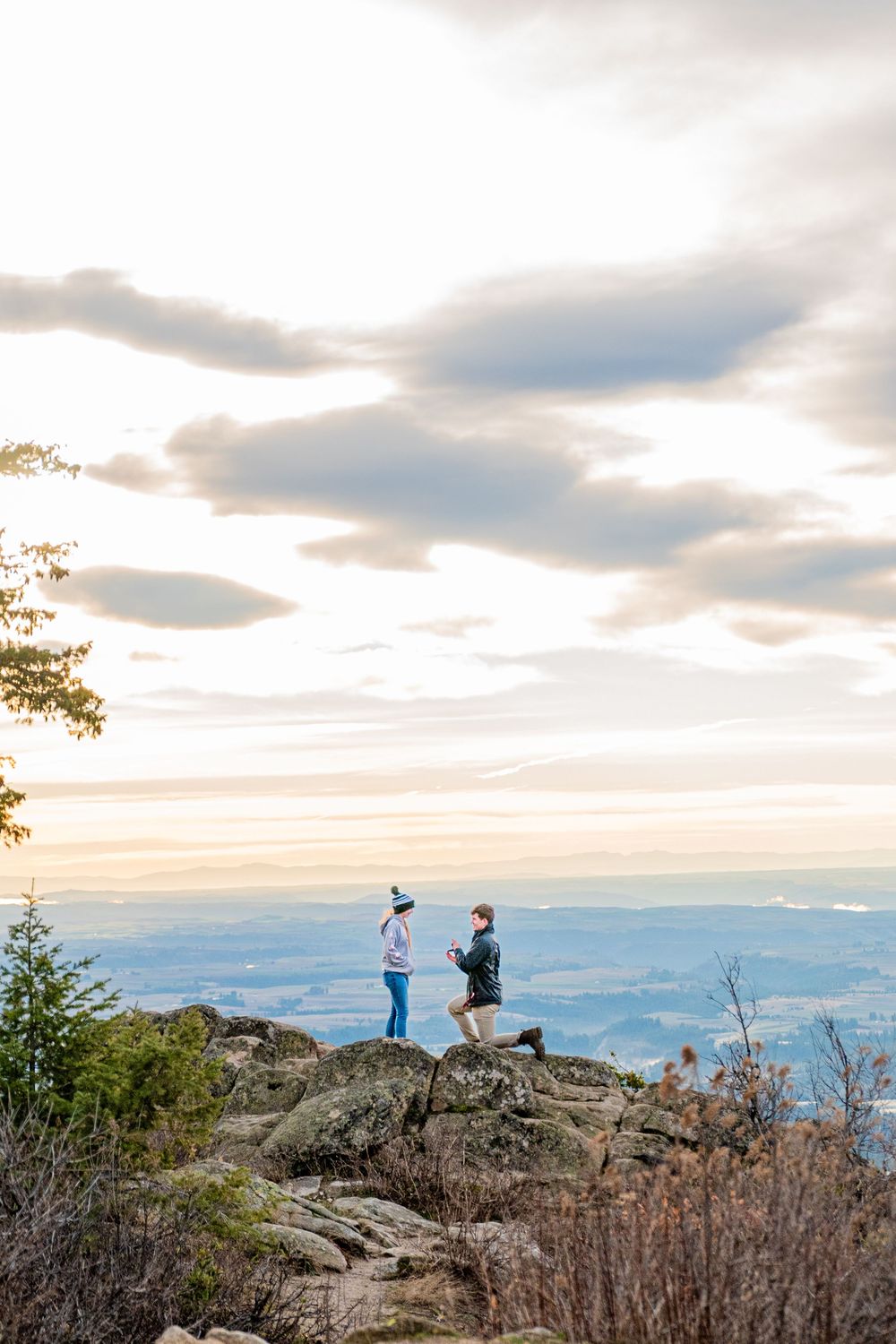 A couple stands on a rocky mountain overlook during sunset with dramatic clouds and valley views in the distance.