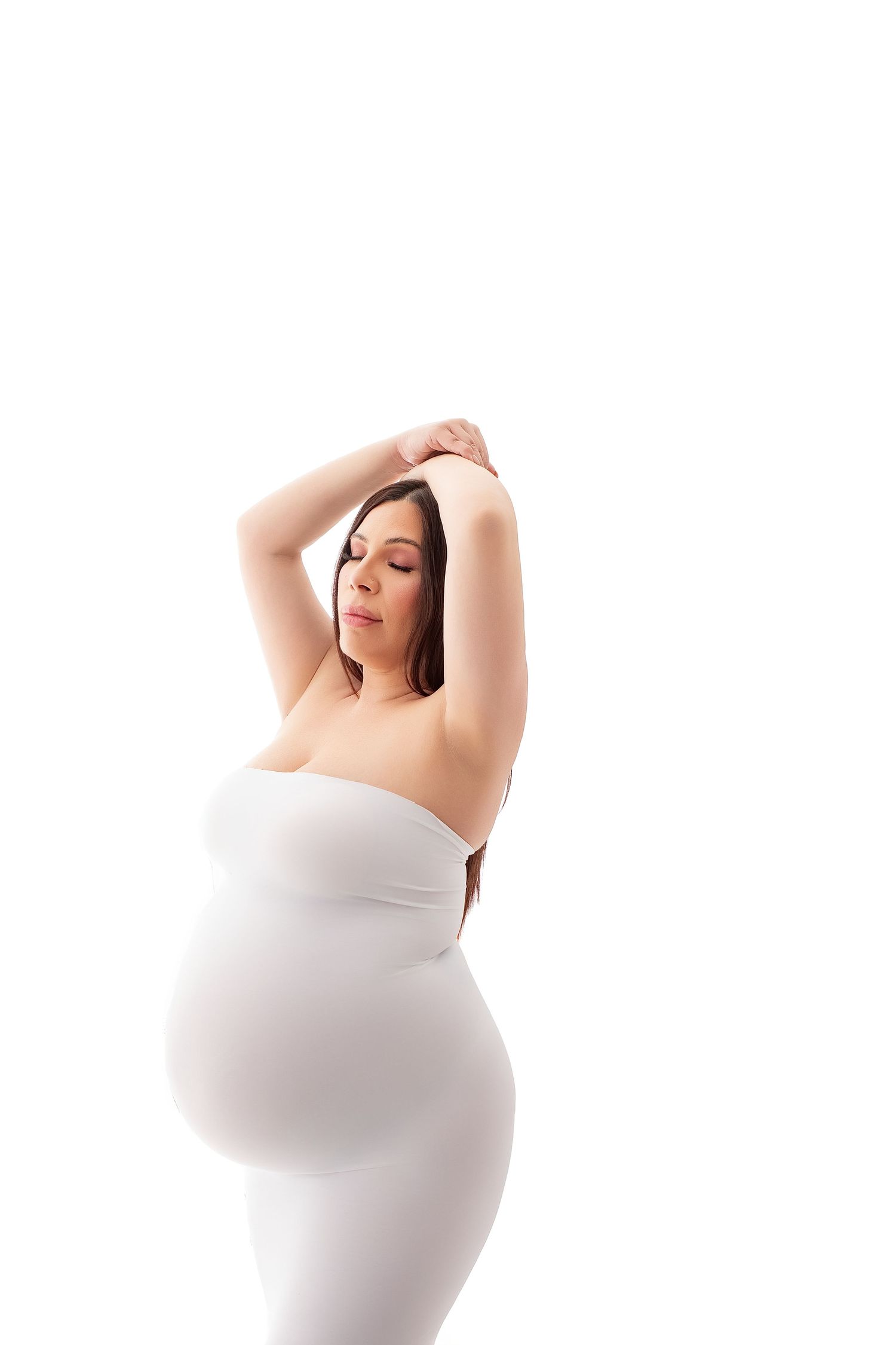 Pregnant woman in white strapless dress with arms raised above head, eyes closed, posing against white background.
