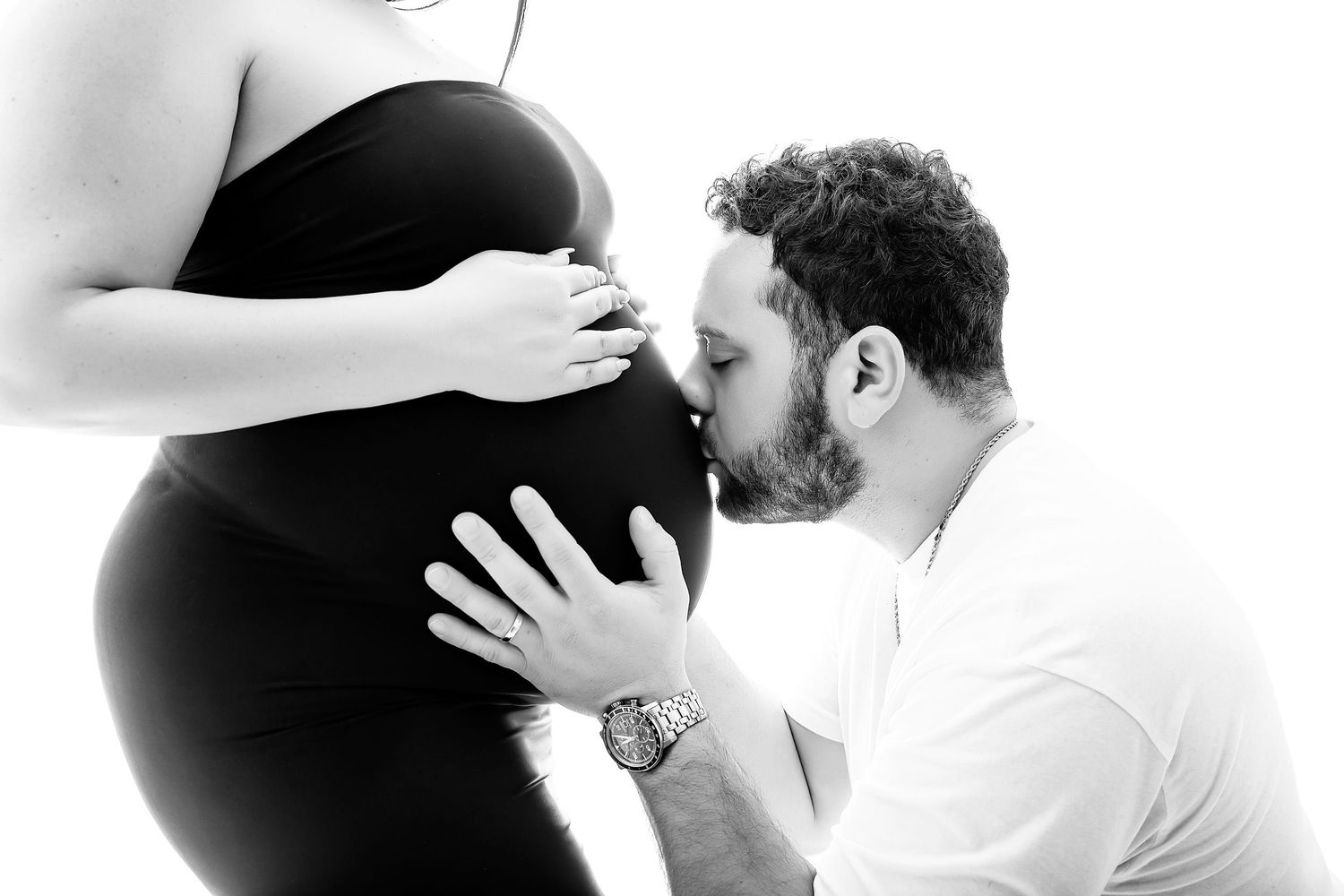 Black and white maternity photo of a man kissing a pregnant woman's belly while she gently holds it.