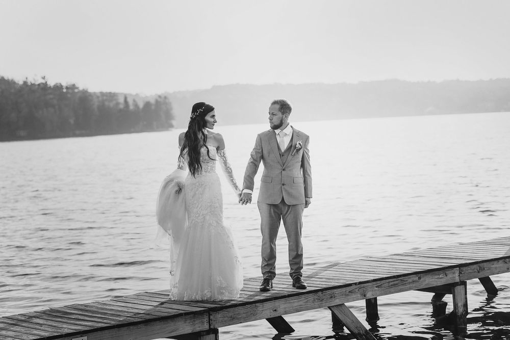 A couple holds hands while standing on a dock overlooking a serene lake in black and white.
