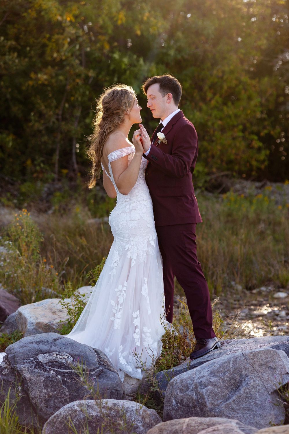 A romantic wedding couple stands on rocks during golden hour in their burgundy suit and lace gown.