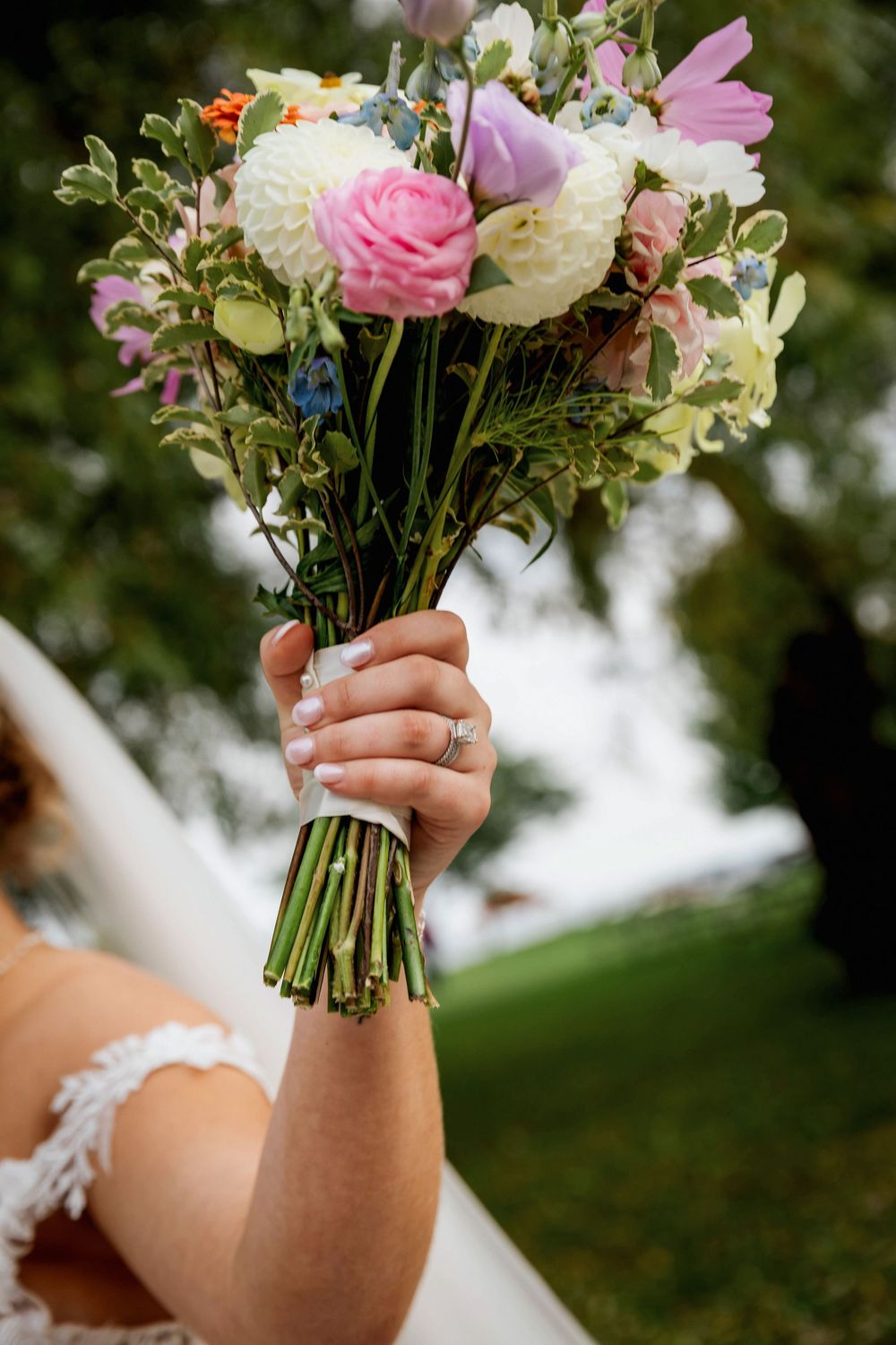 A delicate bridal bouquet with pink and white flowers held up against a green outdoor background.