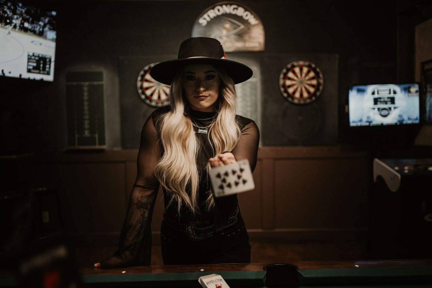 A silhouetted figure in a black hat stands at a bar counter holding playing cards in a dimly lit room with dartboards on the wall.