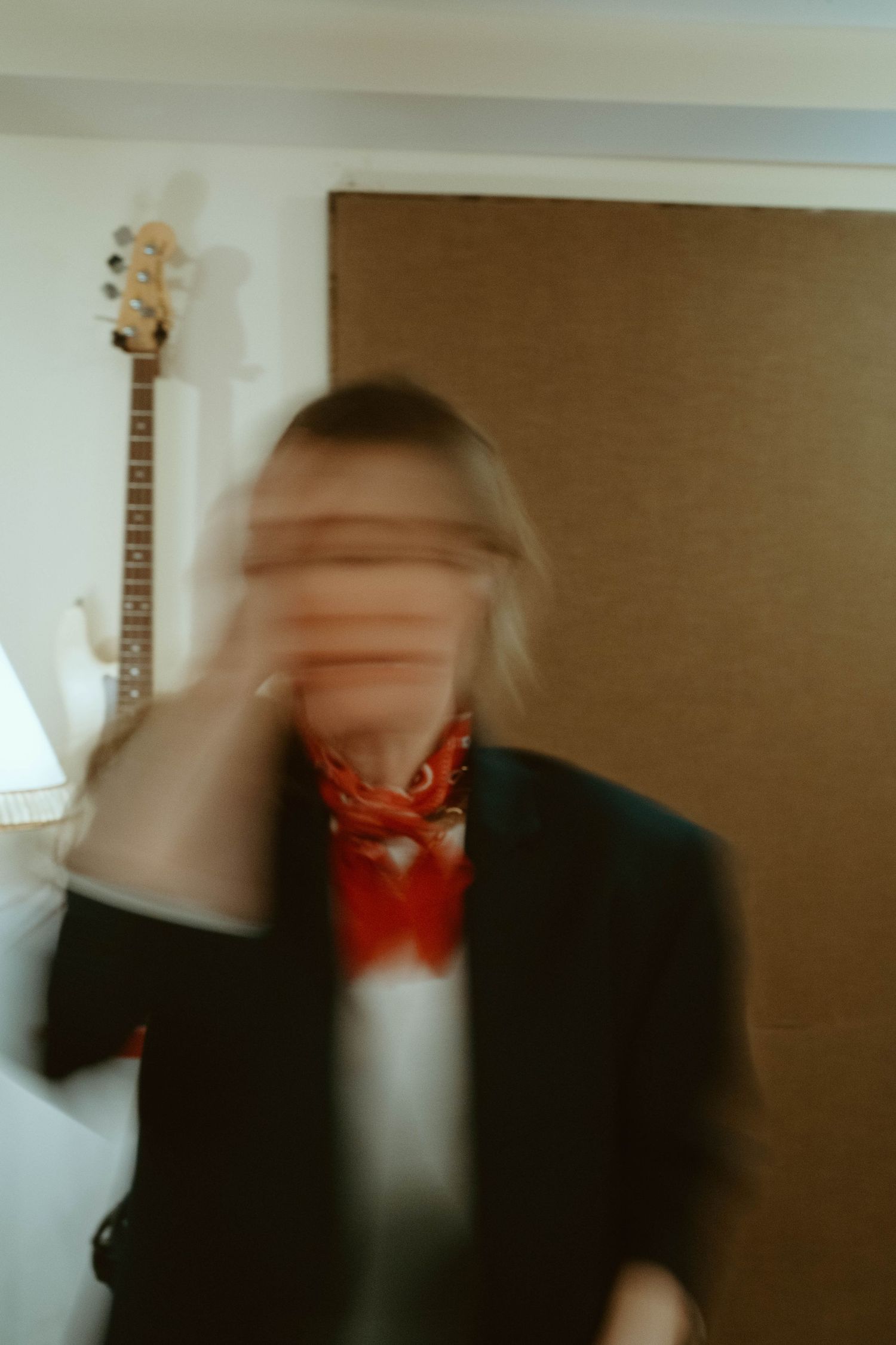 Blurry motion photo of a blonde person in a black blazer and red bandana, with a guitar visible in the background.