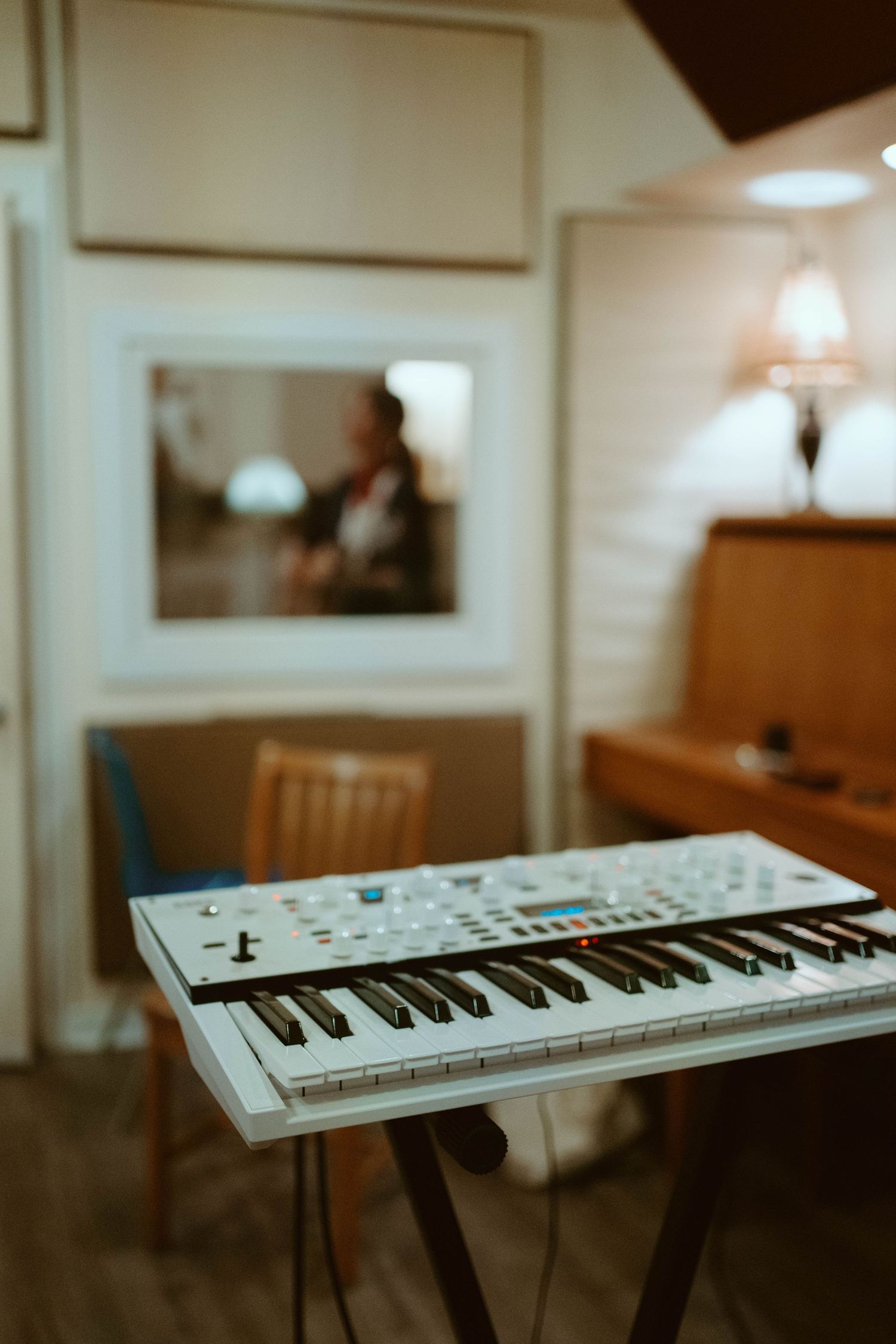 White synthesizer keyboard in foreground with musician visible through studio recording booth window.
