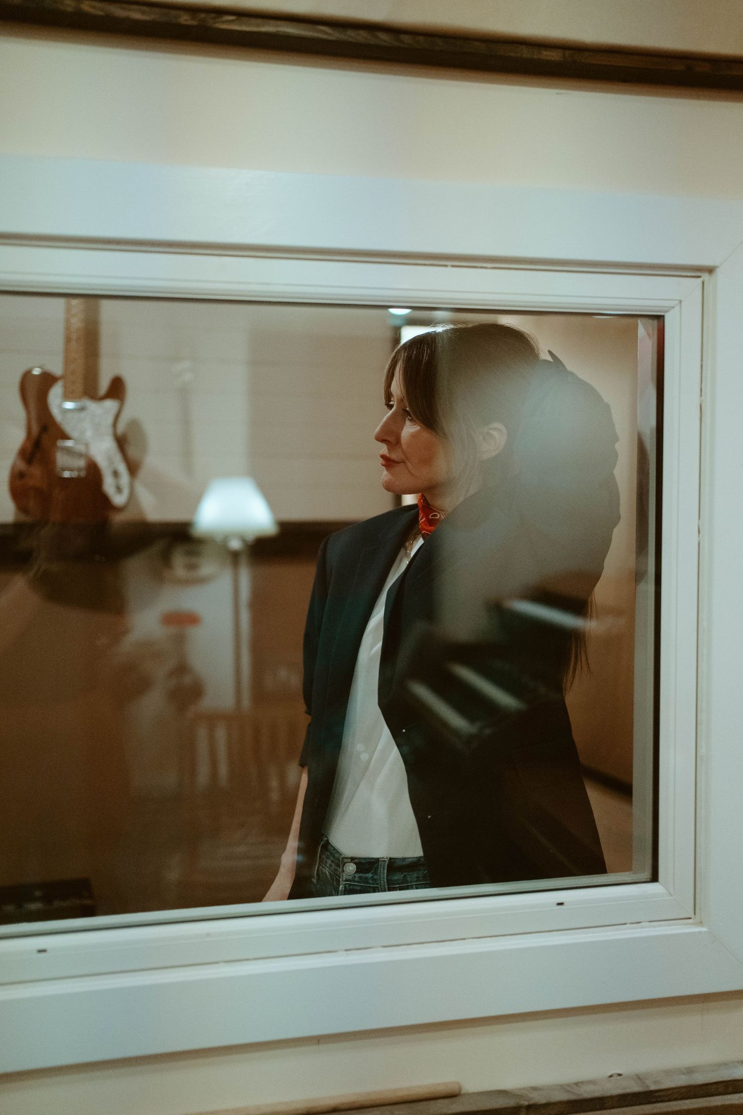 Woman in black blazer and red scarf gazes through a window, with a guitar visible in the reflection behind her.