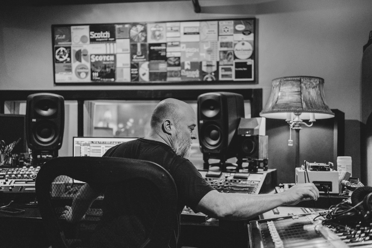Black and white photo of a bald man working at a professional music studio mixing console with monitor speakers.
