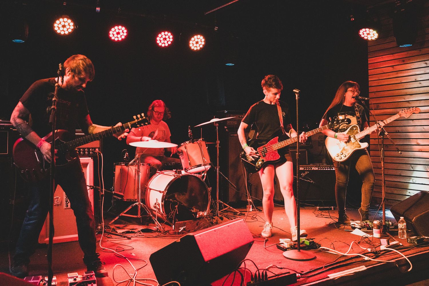 Live band performing on stage with guitars and drums under red and pink stage lights in a dark venue.