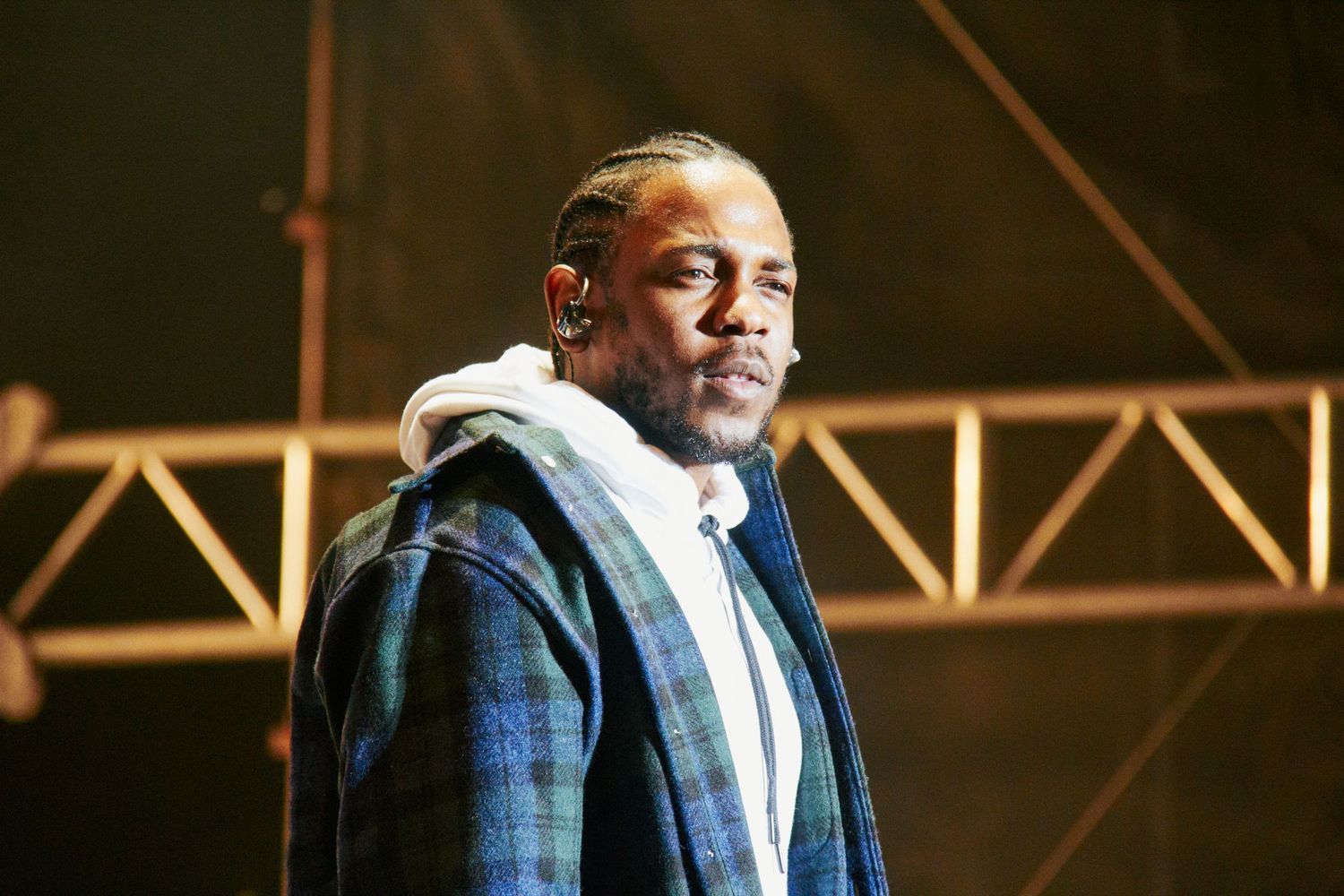 A man in a plaid blazer and white hoodie stands on a stage under metal scaffolding at an outdoor event.