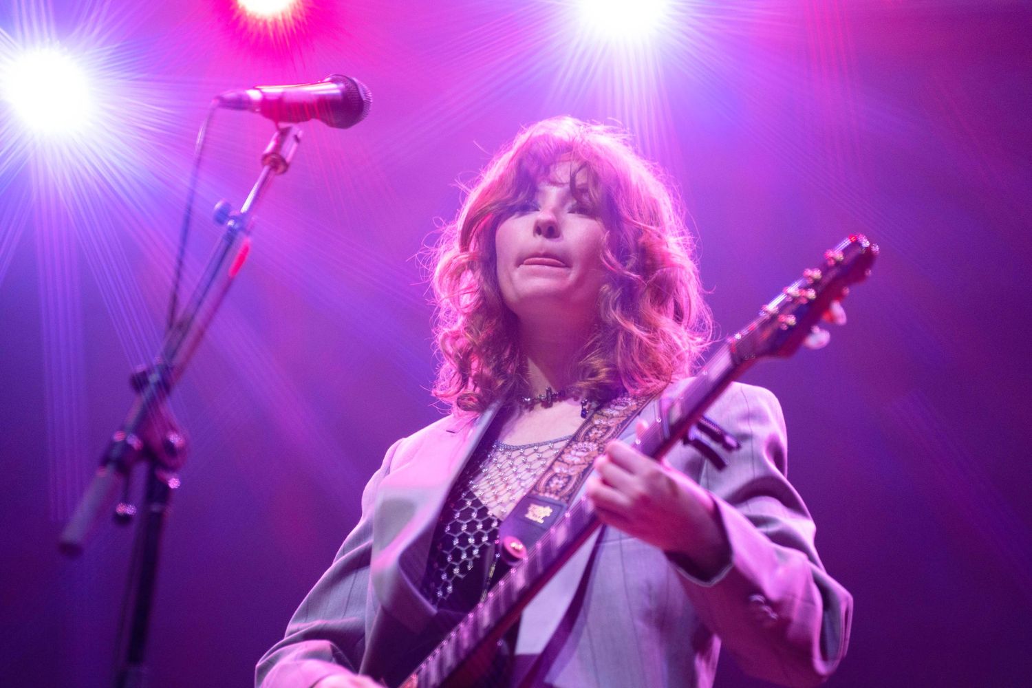 Female guitarist performing on stage under purple lights, playing electric guitar at a live concert.