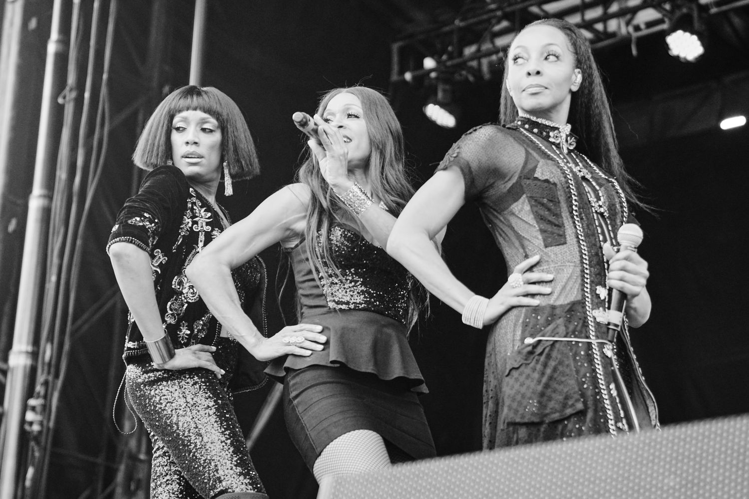 Three women performing on stage in black and white photo, striking poses with hands on hips at outdoor concert.