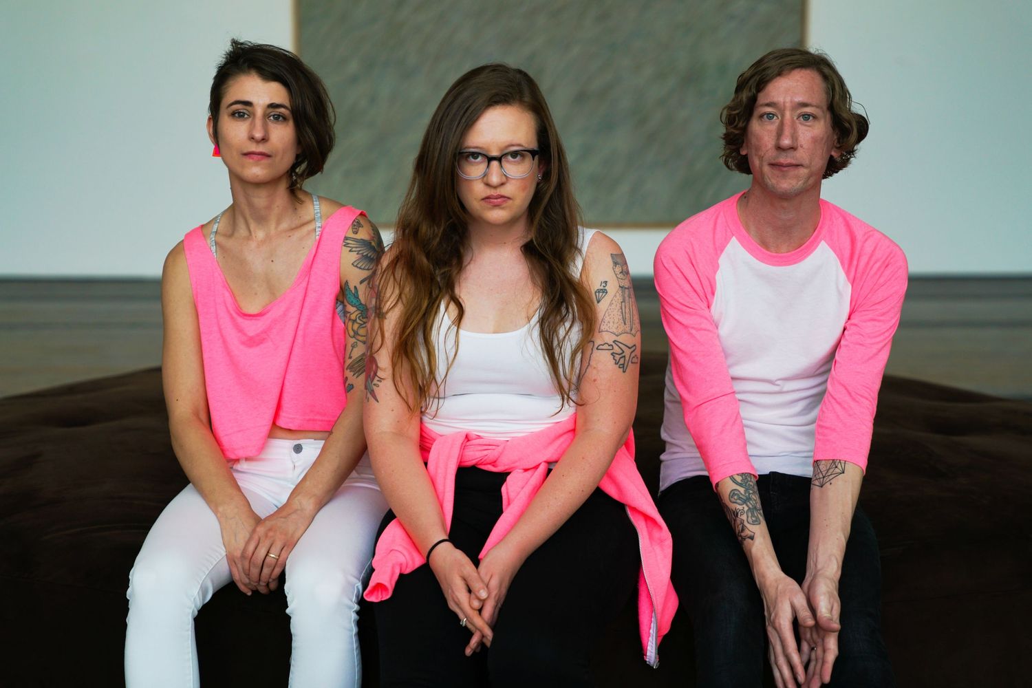 Three people in pink and white outfits with tattoos sitting together against a neutral background.