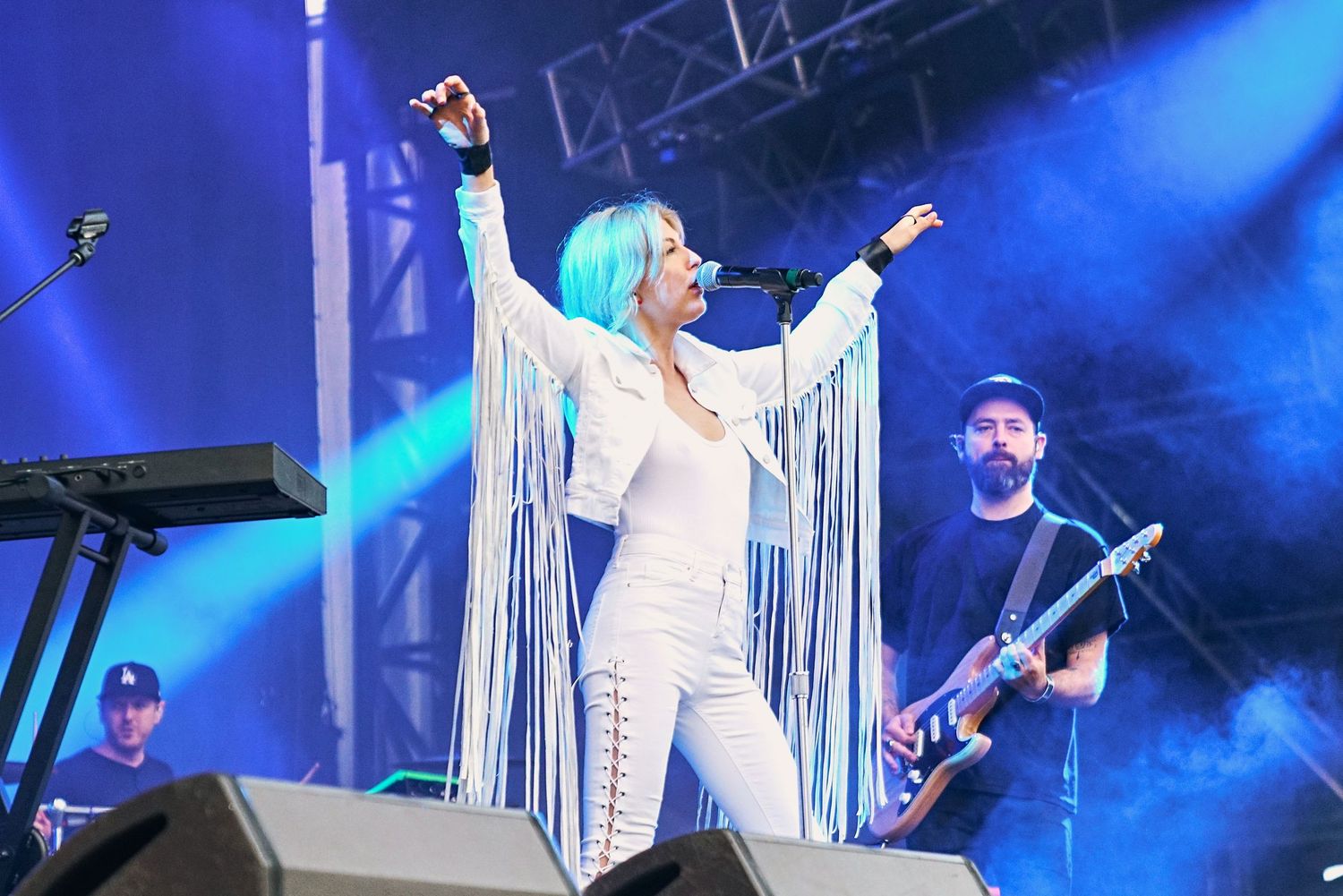 Female singer with blue hair in white fringe outfit performs on stage with band members and keyboards behind her.