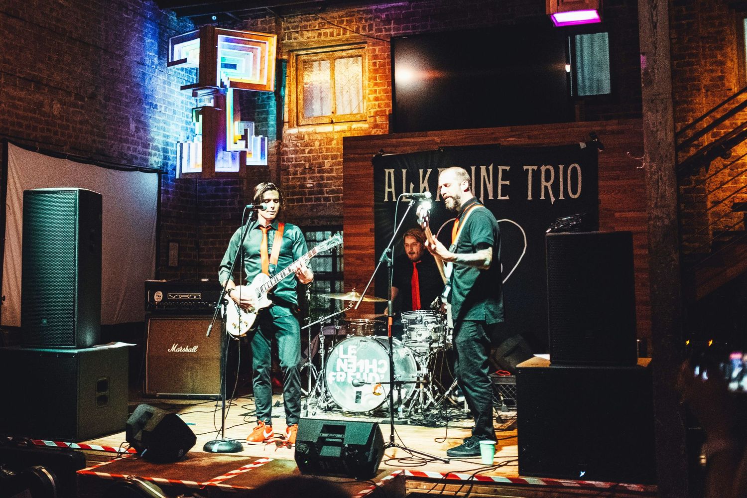 Alkaline Trio performing live on stage at a brick-walled venue with guitarist, drummer, and bassist under colorful lights.